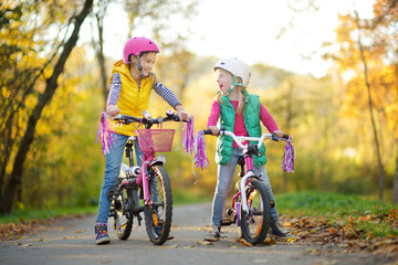 Cute sisters riding bikes in a city park on sunny autumn day. Active family leisure with kids. Children wearing safety hemet while riding a bicycle.