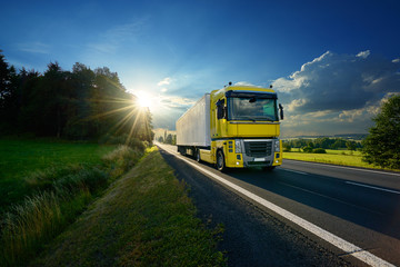 Fototapeta na wymiar Yellow truck arriving on the asphalt road in rural landscape in the rays of the sunset