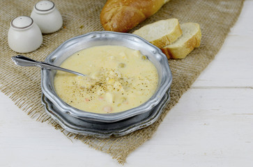 A bowl of hot creamy chicken soup in a rustic setting. Soup is garnished with cracked pepper. Served in a pewter bowl wutg bread and salt and pepper. A spoon rests in the soup.