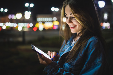 Blogger hipster using in hands gadget mobile phone. Woman pointing finger on blank screen smartphone on background bokeh light in night atmospheric city street.