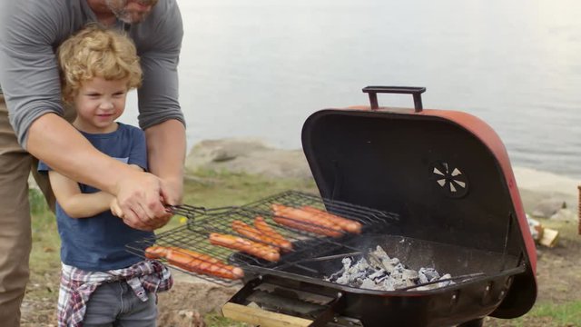 Adorable little boy of elementary school age holding grilling basket and learning to cook sausages, his father helping him