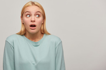 Worried scared blonde girl looks aside around with mouth opened, frightened afraid, presses down...