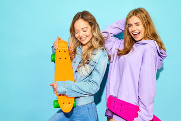 Two young stylish smiling blond women with penny skateboards. Girl in summer hipster sport clothes posing near blue wall in studio. Positive models
