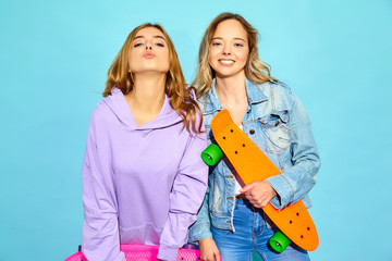 Two young stylish smiling blond women with penny skateboards. Girls in summer hipster sport clothes posing near blue wall in studio. Positive models