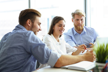 Fototapeta premium Group of young modern people in smart casual wear pointing at laptop and smiling while sitting in the office.