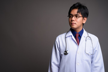 Portrait of young Asian man doctor thinking against gray background