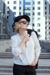 Fototapeta premium Curly blonde with red lips in a black hat on the background of modern buildings