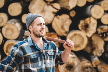 Strong bearded lumberjack wearing plaid shirt hold ax in hand on background of sawmill and warehouse of trees
