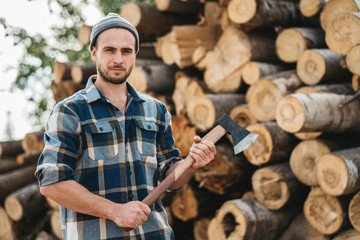 Bearded lumberjack wearing plaid shirt hold ax in hand on background of sawmill