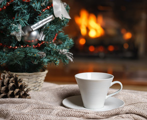 Obraz na płótnie Canvas Cup of tea or coffee, woolen knitted things plaid and Christmas decorations near cozy fireplace background, in country house. Cozy relaxed magical atmosphere home interior. New Year winter concept.
