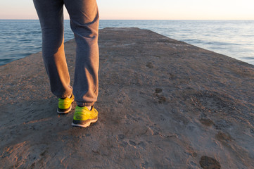 A young man in sneakers is walking along a breakwater to the sea against the background of a sunset