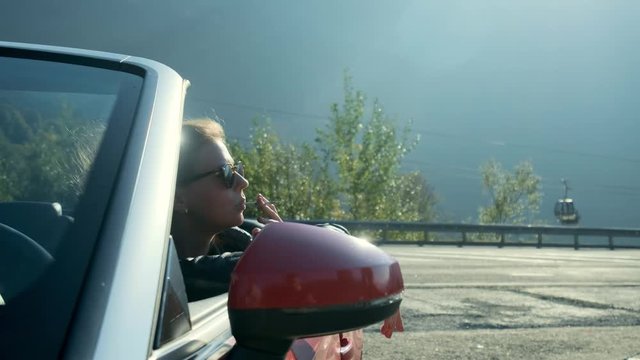 Young girl smoking in a convertible. Young beautiful blonde girl in black glasses and leather jacket smokes in a red cabriolet against the backdrop of high mountains.