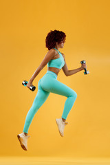 Strong athletic, woman sprinter or runner, running on yellow background with dumbbells wearing sportswear. Fitness and sport motivation.