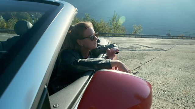 Young girl smoking in a convertible. Young beautiful blonde girl in black glasses and leather jacket smokes in a red cabriolet against the backdrop of high mountains.