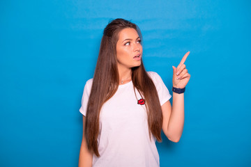Fototapeta premium Cute brunette woman with long hair posing in white t-shirt on a blue background. Emotional portrait. She points her fingers on something, happily