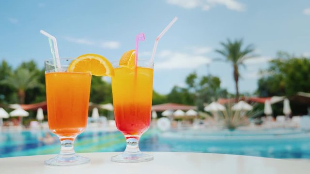 Relax on the sun loungers near the pool with two exotic cocktails with straws, blue sky and green palms in the background near the pool