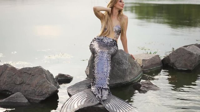 cute attractive mermaid with blond hair and a long silver scaly tail on a stone in the middle of a lake and water, straightens her hair, touches her neck. mysterious picture of a mythical creature