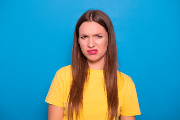 Fototapeta premium Cute brunette woman with long hair posing in yellow t-shirt on a blue background. Emotional portrait. She shows her disgust