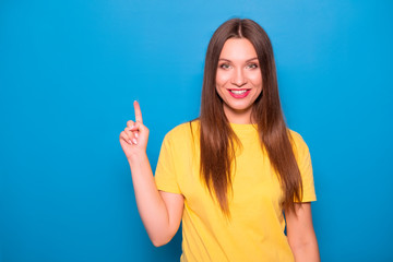 Fototapeta premium Cute brunette woman with long hair posing in yellow t-shirt on a blue background. Emotional portrait. She smiles happily with flawless white teeth and poins finger on something