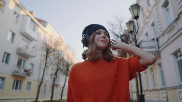 cute young girl walking down old city street and listening music in headphones, urban style, stylish hipster teen in black hat listen music and smile, orange crazy style
