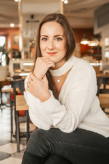 Brunette young caucasian woman in casual sweater and jeans sitting in cafe, smiling happily with a cup of coffee. Lifestyle portrait indoors. Copy space