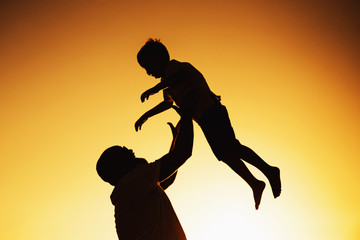 Happy father and son celebrating with him on lap throwing up. Against light on the beach paternal love in father's Day.
