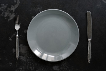 Empty gray plate and vintage fork and knife, black stone background. Top view