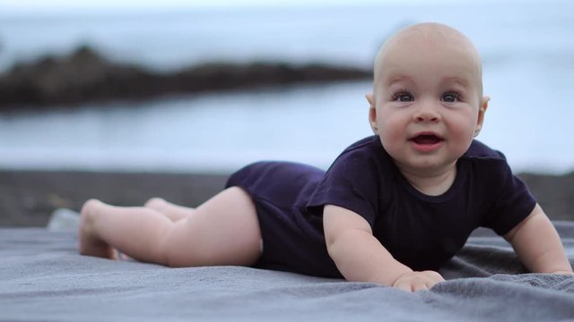 The baby lies on his stomach on the black sand near the ocean and laughs looking at the camera