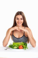 A beautiful smiling girl is sitting at the table with a plate full of fresh vegetables and greens. Studio, white background.