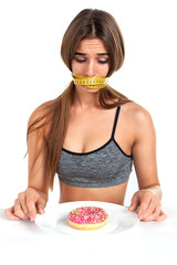Diet concept. Forbidden food. Beautiful slim girl sadly looks at a donut lying on a plate. The girl’s mouth is closed with a measuring tape. Studio, white background.

