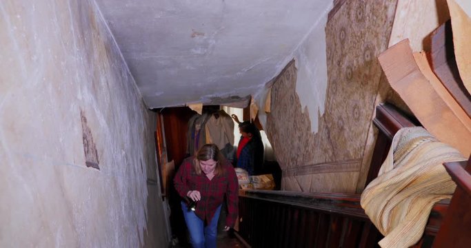 A man and woman walk up the steps of an old abandoned home, looking for antiques and treasures. Wallpaper is falling off the walls.  	