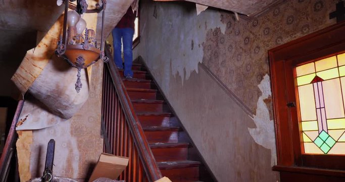 A man and woman walk down the steps of an old abandoned home, looking for antiques and treasures. Wallpaper is falling off the walls.  	
