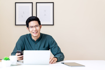 Portrait of young attractive asian businessman or student using mobile phone, laptop, tablet, drinking coffee sitting on desk table wearing green shirt look at camera in home office with copy space.