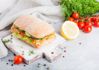 Fresh healthy salmon sandwich with lettuce and cucumber on vintage chopping board on white stone background. Breakfast snack. Fresh tomatoes, dill and lemon.