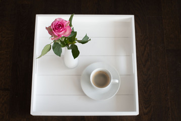 Schwarzer Kaffee in weißer Tasse mit Untertasse neben rosafarbener Rose in weißer Vase auf weißem Tablett von oben