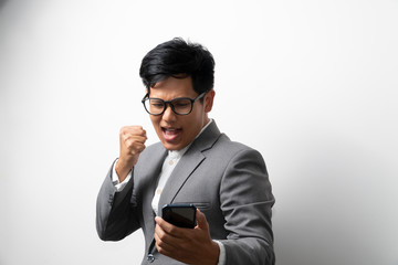 A business man is showing glad and success expression while looking at the smartphone on left hand and make a fist on righthand. More space.