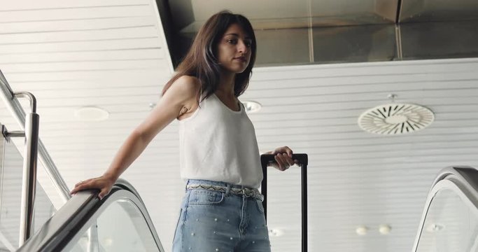 Young beautiful brunette woman goes on escalator in airport with travel bag.