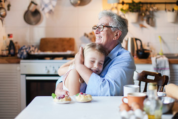 Happy grandmother is hugging granddaughter in cozy home kitchen. Family is cooking together. Senior woman and cute little child girl are smiling. Kid is enjoying kindness, warm hands, care, support. - obrazy, fototapety, plakaty
