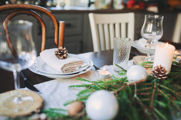 Festive Christmas and New Year table setting in scandinavian style with rustic handmade details in natural and white tones. Dining place decorated with pine cones, branches and candles
