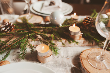 Festive Christmas and New Year table setting in scandinavian style with rustic handmade details in natural and white tones. Dining place decorated with pine cones, branches and candles