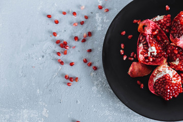 chunks of ripe red pomegranate in black plate with white on light grey, uneven background, close-up