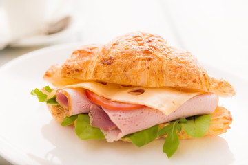 Croissant sandwich with smoked ham, tomato and mixed salad on a white plate.