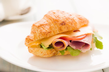 Croissant sandwich with smoked ham, tomato and mixed salad on a white plate.