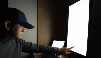 Side view of man Hacker sit at the computer monitor, white screen tablet pointing fingers monitor in the dark room.