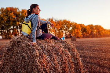 Young traveler having rest in autumn field at sunset. Woman admiring view sitting on haystack