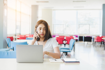 Obraz premium Young asian college woman talking with friends on mobile phone while working with laptop.