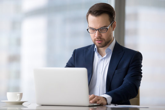 Tensioned or surprised caucasian man is sitting in office and typing on laptop. Serious and concentrated male in suit is working at notebook and looking resent and worried