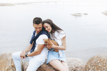 Fototapeta premium Young beautiful positive couple with small dog sitting on stone by sea