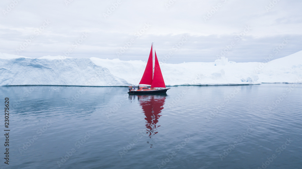 Wall mural beautiful red sailboat in the arctic next to a massive iceberg showing the scale - aerial view as vi