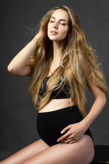 Naklejka premium Beautiful pregnant leggy blonde woman wearing black maternity underwear sits on a chair on a gray background and touches her hair. Health, fashion, motherhood. Commercial design